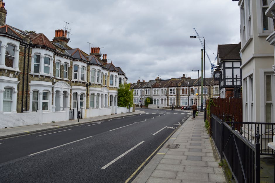A street view of residential houses on a cloudy day, with a sidewalk on the right side featuring a black metal railing and a row of white terraced homes with bay windows and brick detailing. The road 